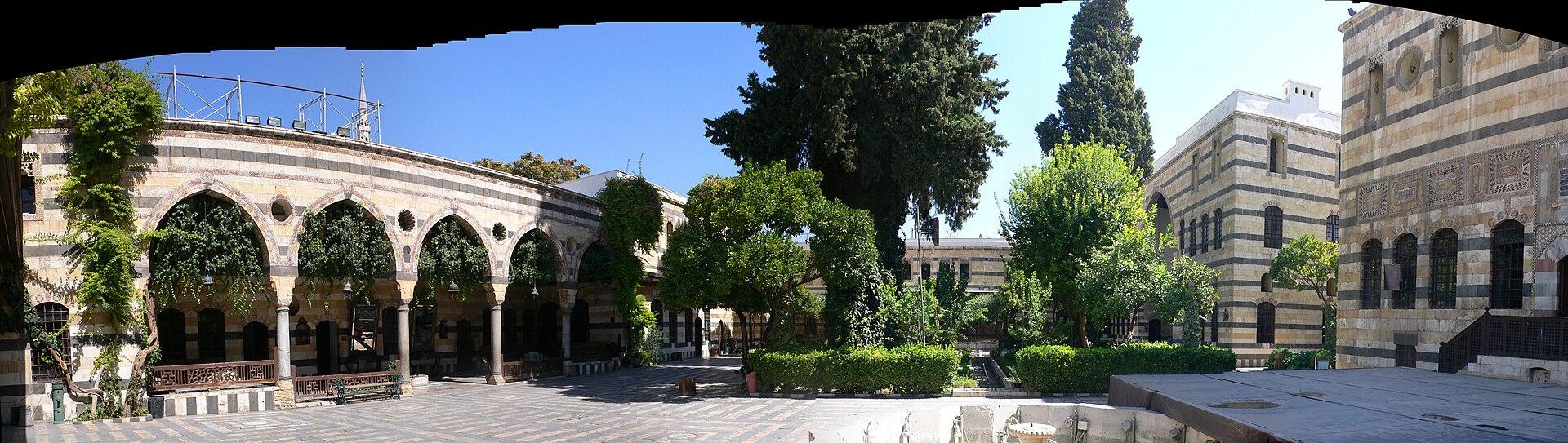 A Damascene courtyard with striped ablaq stonework, an arcaded iwan, citrus trees and a central fountain