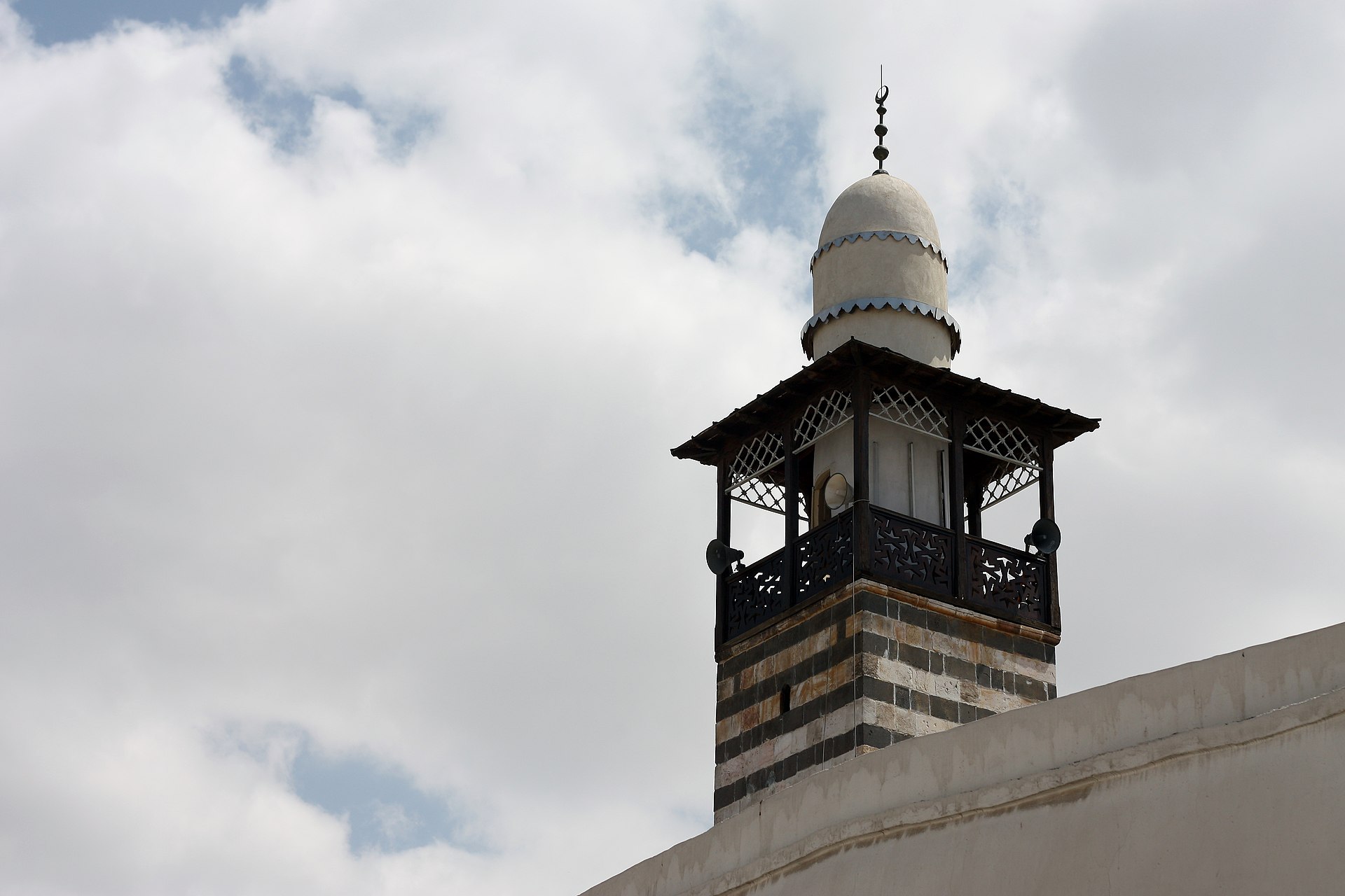 A traditional Damascene minaret in striped stone with a carved wooden gallery, rising above the old city